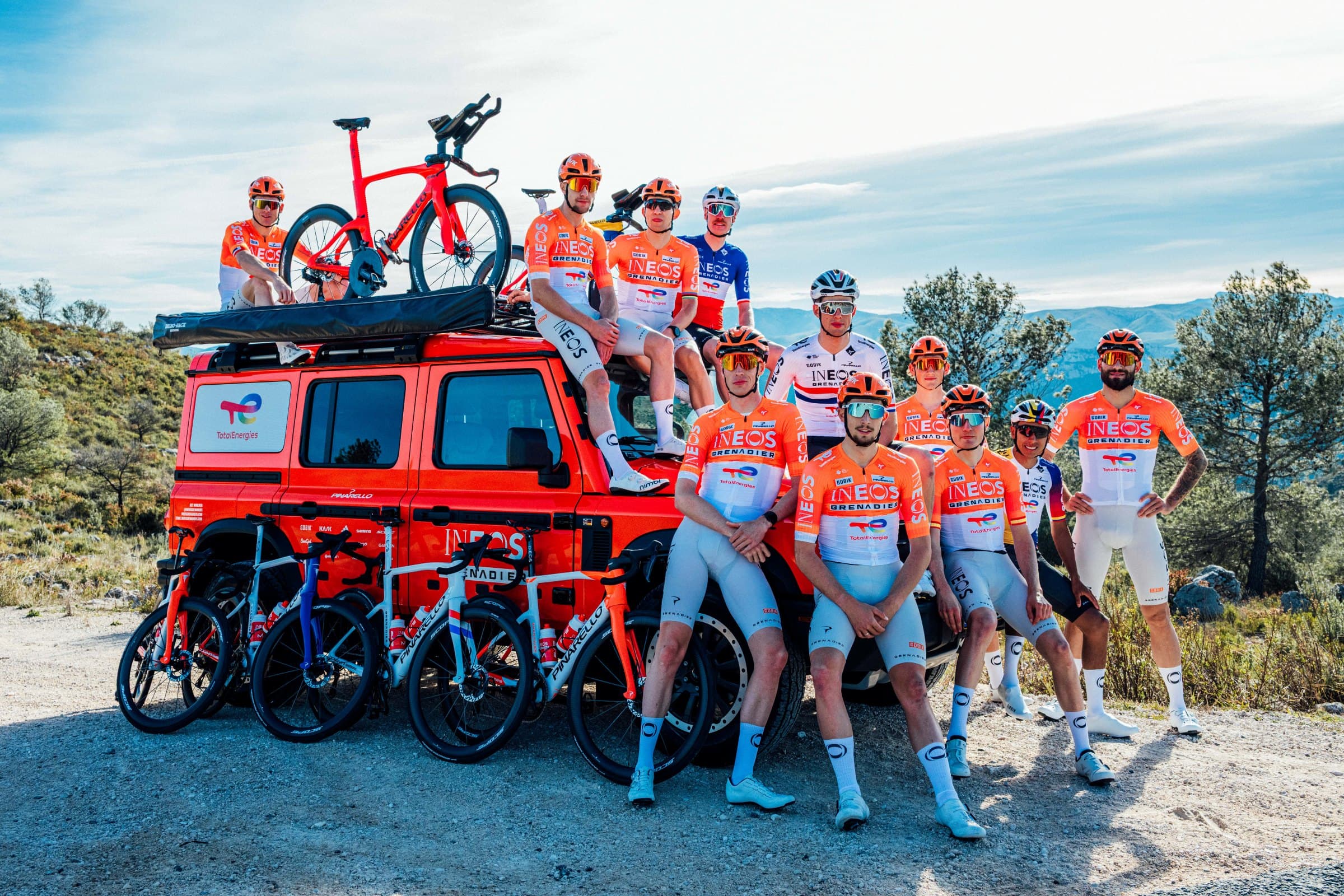 INEOS Grenadiers cycling team in orange and white kit standing with their bikes beside a Grenadier 4x4 during a team appearance.